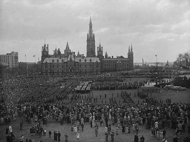 Photographie en noir et blanc – Des milliers de personnes rassemblées près des édifices du Parlement à Ottawa. Une tribune est dressée au milieu de la photo, à droite. Plusieurs véhicules sont visibles dans les environs.